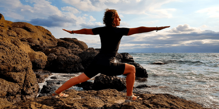 Mulher de preto com roupa de surfe pratica yoga  em meio às pedras do mar no fim de tarde com céu claro. 